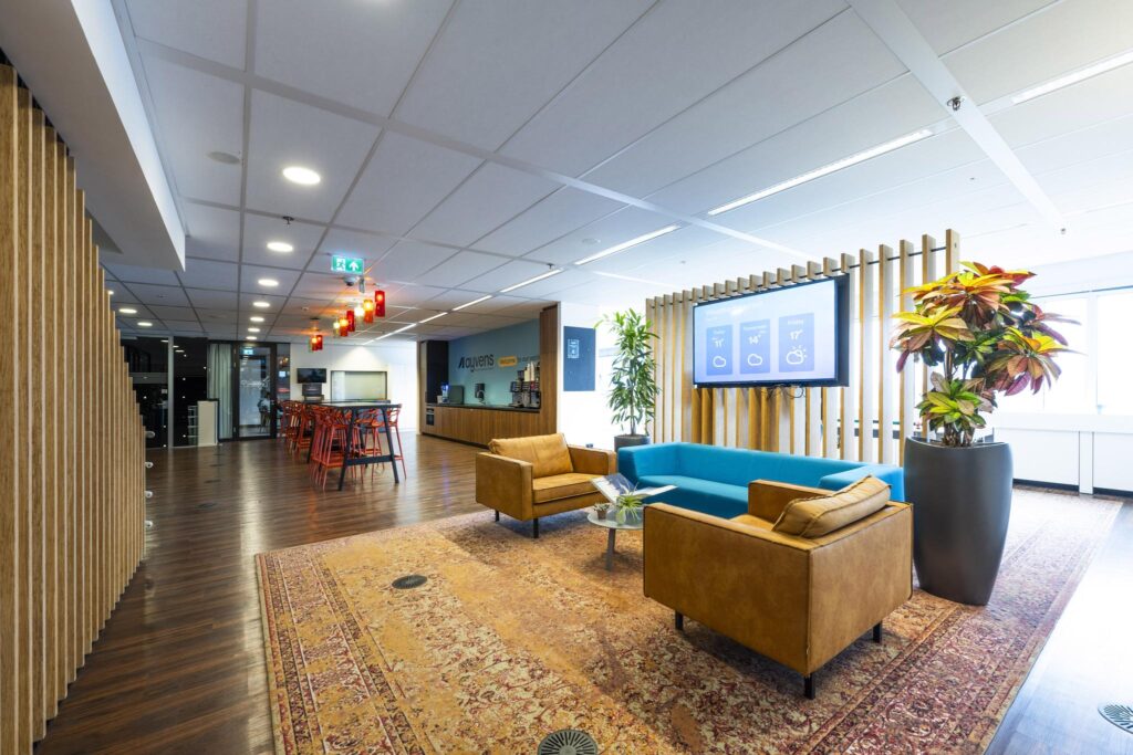 Moderne kantoorlobby aan de Johan Huizingalaan met zitgedeelte, grote planten, flatscreen en een koffiebar met hoge tafels en rode barkrukken.